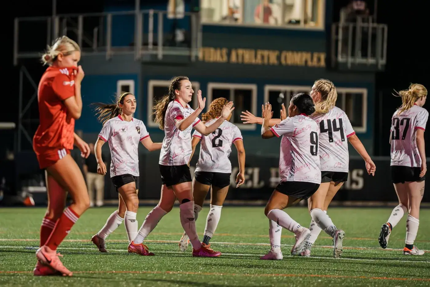 Love City FC players celebrating a goal under the lights at Vidas Athletic Complex